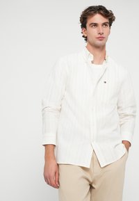 Camicia a maniche lunghe color crema con bottoni, caratterizzata da sottili righe verticali rosse. Tessuto morbido, colletto classico e un piccolo logo sul petto.