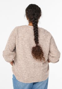 Beżowy sweter wykonany na drutach o luźnym kroju, z opadającymi ramionami i prążkowanymi mankietami. Miękka tekstura i subtelny wzór w cętki. Nosi się go ze niebieskimi dżinsami.