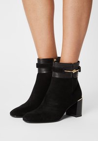 Bottines en daim noir avec un talon épais, un poignet en cuir et des détails en métal doré, offrant un bout pointu et une texture lisse.