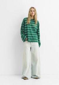 Grønstribet langærmet skjorte med gul tekst, parret med vide lyse denimjeans. Casual pasform, blødt stof og afslappet silhuet.
