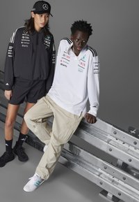 Black athletic jacket with logo accents paired with black shorts. White long-sleeve shirt features logo prints, beige cargo pants. Urban setting.