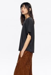 Camiseta negra de manga corta con un corte holgado, que presenta detalles de tachuelas metálicas en los hombros. Combinada con pantalones de ante marrón de pierna ancha.