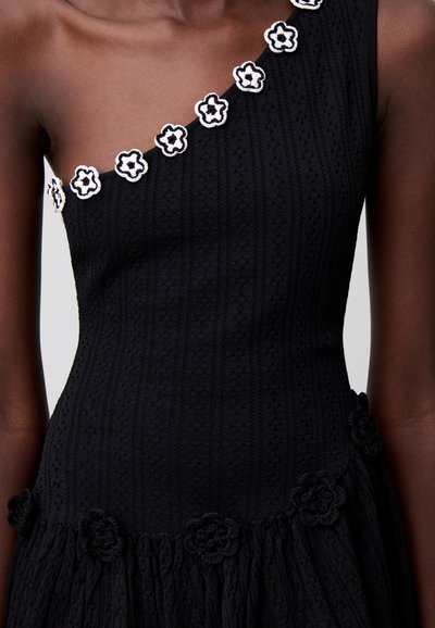 Primo piano di una persona dalla pelle scura che indossa un vestito nero monospalla con decorazioni floreali bianche e nere lungo lo scollo e l'orlo.
