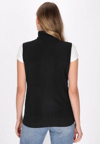 Schwarze ärmellose Fleece-Weste mit hohem Kragen, glatter Textur und klaren Linien. Über einem weißen Hemd getragen, kombiniert mit blauen Jeans.