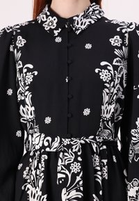 Μαύρο φόρεμα με λευκό λουλουδάτο σχέδιο, κουμπιά μπροστά, γιακά και ζώνη στη μέση. Μακριά μανίκια με συγκεντρωμένες λεπτομέρειες και μαλακή υφή υφάσματος.