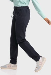 Marineblaue Hose mit einem elastischen Bund, ausgestattet mit einem Kordelzug und Seitentaschen, kombiniert mit hellen Sneakers, die einen lässigen Look unterstreichen.