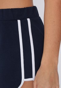 Pantaloni sportivi blu con piping bianco ai lati, vita elasticizzata e una texture morbida; mostra parte di una mano contro il tessuto di lato.