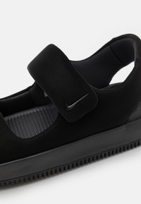 Chaussure noire dotée d'une tige en cuir texturé, d'une sangle Velcro et d'une semelle nervurée pour une meilleure adhérence. Le logo Nike est visible sur la sangle.