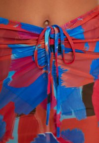 Taille et nombril visibles avec un tissu tie-dye rouge vif et bleu noué en nœud autour de la taille.