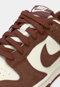 Karakana smešane smeđe i krem Nike patike izbliza, prikazujući teksturirane vezice, materijal od antilopa i Nike swoosh logo sa strane i na jeziku.