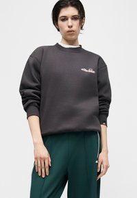 Άτομο που φοράει σκουρόγρινο φούτερ ellesse και σκούρο πράσινο παντελόνι με λευκές ρίγες στα πλάγια, στέκεται μπροστά σε ένα απλό φωτεινό φόντο.