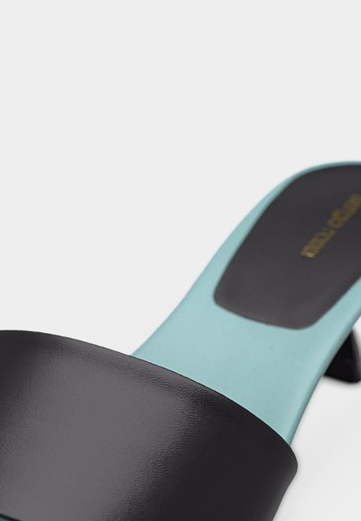 Κοντινό πλάνο σε μαύρο σανδάλι τύπου σλάιντ με φαρδιά λουρίδα και ανοιχτό μπλε πέλμα, με χαμηλό τακούνι και χρυσό λογότυπο της μάρκας στην εσωτερική σόλα.