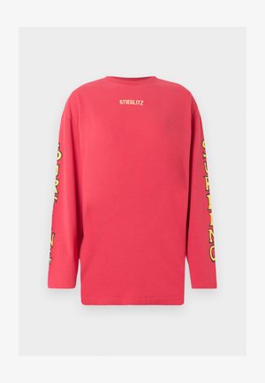 Κόκκινο πουκάμισο με μακριά μανίκια με την ένδειξη «STIEGLITZ» σε μικρό γράμμα στο επάνω μέρος της πλάτης και κίτρινο στιλιζαρισμένο κείμενο που τρέχει κατά μήκος και των δύο μανικιών.