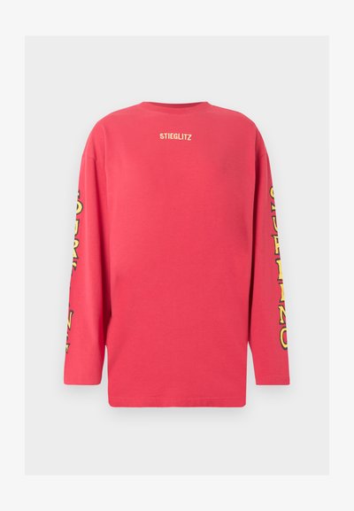Κόκκινο πουκάμισο με μακριά μανίκια με την ένδειξη «STIEGLITZ» σε μικρό γράμμα στο επάνω μέρος της πλάτης και κίτρινο στιλιζαρισμένο κείμενο που τρέχει κατά μήκος και των δύο μανικιών.