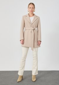 Bež villasegu mantel, millel on vöökoht, lõhiklapid, ühe nööbi sulgemine ja pikad varrukad, koos kreemjate pükste ja beezide kontsakingadega.