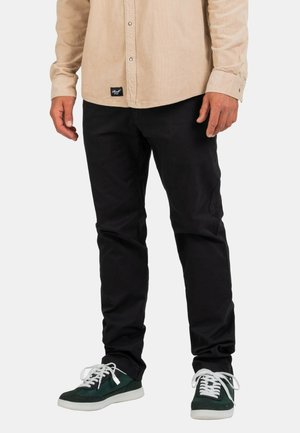 Schwarze Baumwollhose mit schmaler Passform, kombiniert mit grün-weißen Sneakers. Beigefarbenes Cordhemd mit Knöpfen.