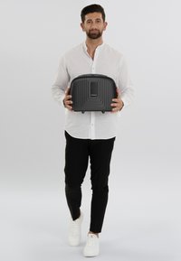 Zwarte harde tas met ribbeltextuur, een handvat aan de bovenkant en een voorvak. Het model houdt het tegen een lichte achtergrond.