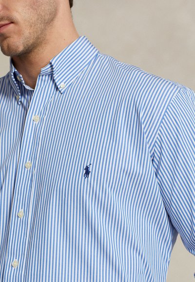 Mann iført en blå og hvit vertikalt stripet skjorte med knapper og en liten brodert marineblå polo-spillerlogo på brystet.