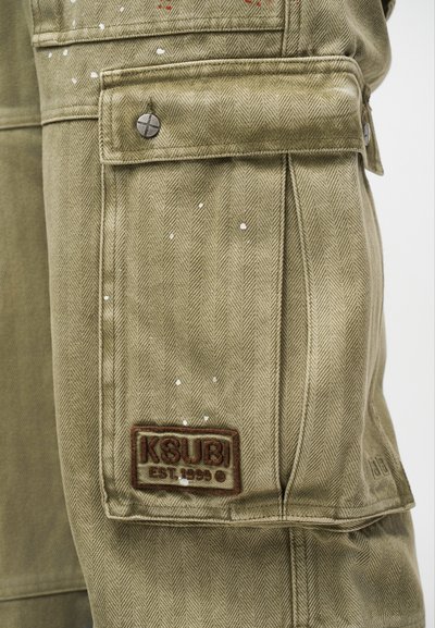 Κοντινό πλάνο της τσέπης cargo από ύφασμα καμηλό με σχέδιο ψαροκόκαλο, μεταλλικό κουμπί με σφραγίδα και μπαλώμα "KSUBI EST. 1999", με μικρές λευκές πιτσιλιές από μπογιά.