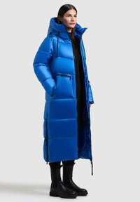 Langer blauer Puffer-Mantel mit Kapuze, der ein gestepptes Design, Reißverschlusstaschen und eine glänzende Oberfläche aufweist. Kombiniert mit schwarzen kniehohen Stiefeln.