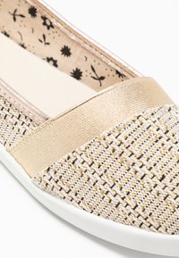 Chaussure plate à enfiler avec un dessus en tissu tissé beige et doré, présentant une bande d'accent dorée et une semelle intérieure à motif floral.