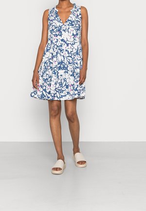 Femme portant une robe bleu sans manches à fleurs blanches et roses, associée à des sandales beige ouvertes à l'avant, debout sur un sol gris.
