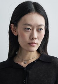 Sølv halskjede med en liten båtanheng på en tynn kjede, båret over en svart, knappet, gjennomsiktig skjorte med krage. Glatt tekstur.