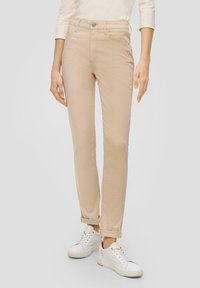 Pantalones slim-fit de color beige claro, confeccionados con una tela suave, que cuentan con cinco bolsillos, cierre de botón y puños enrollados. Llevados con zapatillas blancas.
