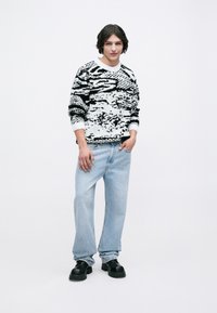 Suéter de patrón en blanco y negro, jeans de tiro relajado en azul claro y zapatos gruesos negros. El suéter tiene mangas largas y un escote redondo.