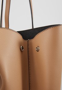 Sac fourre-tout en cuir marron clair avec intérieur noir, doté d'une fermeture à bouton-pression argentée et de deux bandoulières.