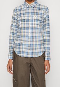 Ljusblå rutig skjorta med gula, gröna och rosa ränder, knapplist framtill, två bröstfickor och rundad nederkant.