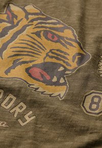 Un tejido verde presenta un gran gráfico de un tigre en amarillo y marrón con acentos rojos, acompañado de textos y diseños numéricos en un color claro.