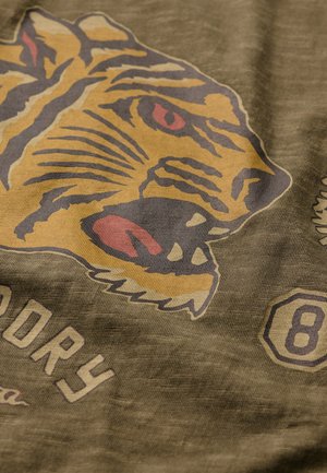 Grønt stof har et stort tiger grafik i gul og brun med røde accenter, ledsaget af tekst og numeriske design i lys farve.