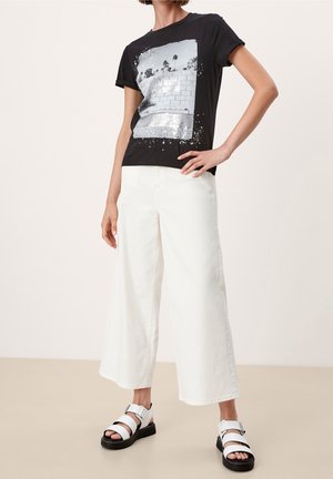 Person iført sort t-shirt med grafik, hvide bukser med vide ben og hvide sandaler med sorte såler, stående med hånden på hoften.