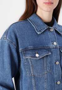 Denim jakna v srednji modri barvi s klasičnim ovratnikom, dvema prsnikoma, srebrnimi gumbi in vidnimi šivi. Teksturiran material.