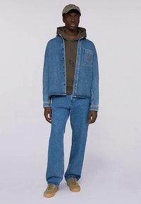 Jeansjacka och byxor i ljusblått med en fickdetalj, kombinerat med en mörkgrön hoodie, beige sneakers och en keps.