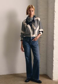 Overhemd met knopen en geometrische strepen in marineblauw en crème, gecombineerd met middelhoge blauwe jeans en zwarte schoenen. Sjaal geknoopt bij de kraag.