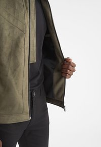 Olive green suede jacket with a smooth interior, zip closure, and a textured exterior featuring diagonal stitching. Inside is a black lining.