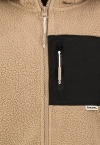 Braune Fleecejacke mit strukturiertem Oberteil, schwarzer Reißverschlusstasche auf der Brust und silbernem Zubehör. Verfügt über einen Frontreißverschluss und eine verstellbare Kordel.