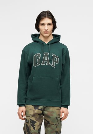 Joven con sudadera con capucha GAP de color verde oscuro y pantalones cargo de camuflaje, de pie mirando hacia adelante con expresión neutral sobre un fondo liso.