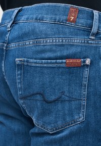 Πίσω τσέπη μπλε τζιν με κόκκινα ετικέτες μάρκας, αντίθετη ραφή και ένα διακριτικό κεντητό σχέδιο κύματος.