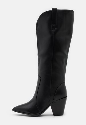 Mustat nahkaiset polven yli ulottuvat saappaat, joissa on terävä kärki ja kapeneva korko. Sileä pinta, minimalistinen muotoilu ja sivuompelut. 