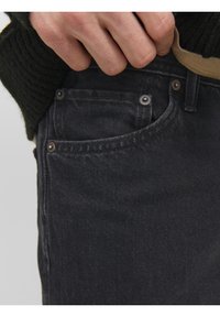 Svarta jeans i denim med en närbild av framfickan och nitar. Tyget har en mjuk textur och en subtil lyster.