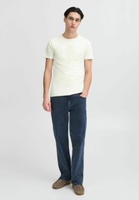 Tricou galben deschis cu guler rotund, purtat cu blugi denim de culoare bleu închis. Blugii au o croială lejeră, iar modelul poartă papuci maro.