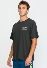 T-shirt en coton noir avec un col rond, des manches courtes, et un logo Volcom blanc sur la poitrine gauche. Porté avec un short bleu à motif géométrique.