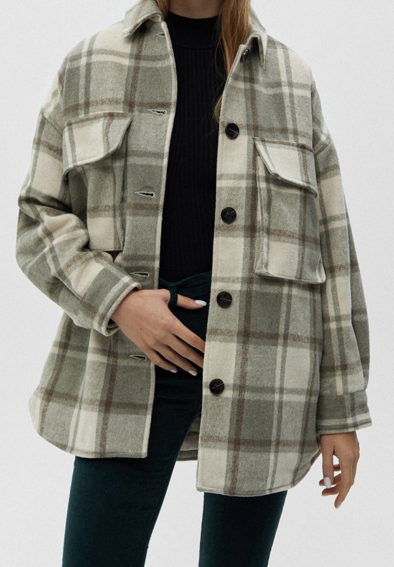 Femme portant un manteau oversize à carreaux beige et gris avec des boutons noirs, associé à un haut noir et un pantalon foncé, main posée sur la taille.