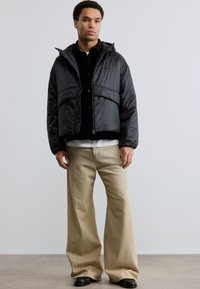 Άνδρας που στέκεται, φορώντας μαύρο μπουφάν τύπου puffer, μαύρο πουκάμισο, λευκό εσώρουχο, μπεζ παντελόνι με φαρδιά μπατζάκια και μαύρα παπούτσια μπροστά σε απλό φόντο.