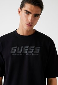Must musta T-särk, millel on peegeldav "GUESS" logo ja all tekst "EST.1981 - CALIFORNIA", ümmargune kaelus ja lühikesed varrukad.