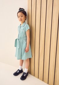 Groen-wit geruit jurkje met korte mouwen, een kraag en een ceintuur in de taille. Zwarte lakleren schoenen en witte sokken maken de outfit compleet.