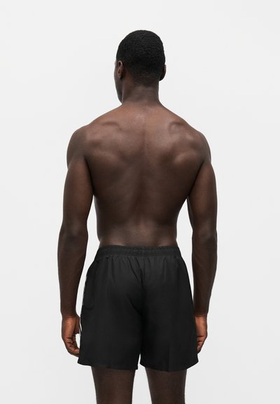 Homme musclé debout, dos à la caméra, portant un short de sport noir sur un fond blanc uni.
